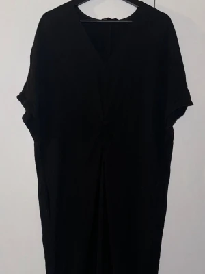 Svart långklänning med fransar från Zara - Svart långklänning från Zara med v-ringad hals och korta ärmar. Klänningen har snygg knutdetalj framtill och långa fransar nedtill som ger en cool touch. Tillverkad i ett mjukt material som känns skönt mot huden. Perfekt för dig som gillar stilrena och trendiga plagg.