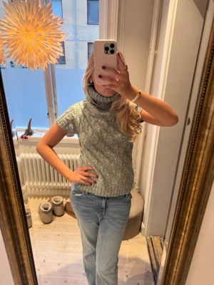 Grå stickad polotröja med kort ärm - Säljer en grå stickad polotröja med korta ärmar och snyggt flätmönster. Tröjan har en hög och bred polokrage som ger extra mysfaktor. Passformen är normal och materialet känns mjukt och värmande. Perfekt att styla med jeans eller kjol.