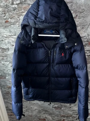 Mörkblå Polo Ralph Lauren jacka - Säljer en mörkblå pufferjacka från Polo Ralph Lauren med huva och dragkedja framtill. Jackan har två sidofickor med dragkedja och ett litet rött Polo-logo broderat på bröstet. Perfekt för kalla dagar och har en schysst, sportig look.