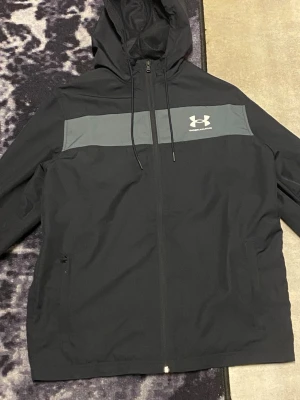 Svart vindjacka från Under Armour - Svart vindjacka från Under Armour med grått band över bröstet och logga. Jackan har huva med snörning, dragkedja framtill och två sidofickor. Materialet är lätt och syntetiskt, perfekt för blåsiga dagar. Passformen är loose och jackan är långärmad.