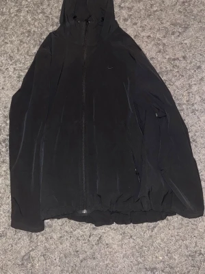 Svart vindjacka från Nike - Svart vindjacka från Nike med huva och dragkedja framtill. Jackan har en minimalistisk design med diskret Nike-logga på bröstet och justerbar nederkant. Perfekt för dig som gillar sportig och clean stil. Den är i storlek L men passar även M väldigt bra. Pris kan diskuteras vid snabb affär. Den är köpt hos JD och är knappt använd.