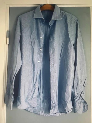 Randig blå skjorta från J.Lindeberg - Säljer en stilren blå skjorta från J.Lindeberg med tunna vita ränder. Skjortan har klassisk krage, långa ärmar och knäppning framtill. Perfekt för dig som gillar en clean och tidlös look. Materialet är mjukt och känns skönt mot huden.