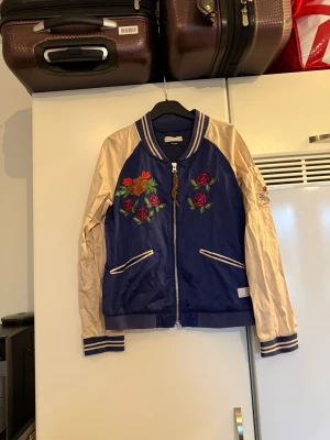 Blå bomberjacka Odd Molly med broderier - Snygg blå bomberjacka från Odd Molly med beige ärmar och broderade blommor framtill samt en stor orange fjäril på ryggen. Jackan har dragkedja, randiga muddar och detaljerade fickor. Perfekt för dig som vill sticka ut med coola broderier och sportig stil. Från märket Odd Molly. Storlek 2 som enligt deras storleksguide motsvarar en M. Mer använd men otroligt vacker skick. 