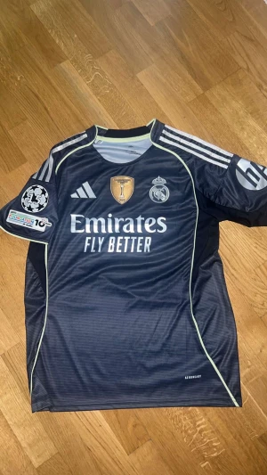 Real Madrid Bellingham #5 Champions League tröja - Säljer en mörkblå Real Madrid Champions League matchtröja med Bellingham och nummer 5 på ryggen. Tröjan har silverfärgade Adidas-ränder på axlarna, broderade klubbmärken och Champions League-patch på ärmen. Materialet är lätt polyester med AEROREADY-teknologi.