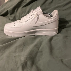 Nike Air Force 1 helvita sneakers  - Klassiska Nike Air Force 1 sneakers i helvitt skinn med låg profil och rund tå. Skorna har perforerad tåbox, snörning och den ikoniska swoosh-loggan på sidorna. Perfekta för en clean och stilren look.