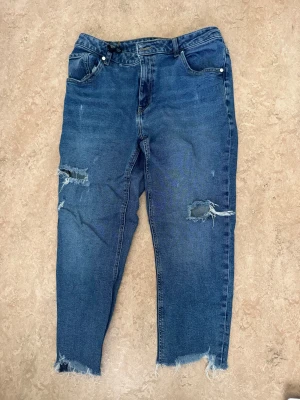 Blå croppade jeans från Lindex - Säljer ett par blå jeans från Lindex med croppad passform och råa, fransiga benslut. Jeansen har slitna detaljer och hål på båda benen för en edgy look. Klassisk femficksmodell och midwaist. Perfekt för dig som gillar streetstyle.