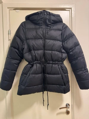Svart pufferjacka från Everest - Säljer en svart pufferjacka från Everest med huva och snörning i midjan. Jackan har långa ärmar, dragkedja framtill och vita detaljer med Everest-tryck på sidorna. Perfekt för kalla dagar och värmer jättemycket. Köpte den för cirka 3 veckort sen och är helt oanvänd. Säljer den då jag känner att  jag inte har någon användning för den. Orginalpris 1000 kr, säljer den för 900 kr men kan tänka mig gå ner i pris☺️