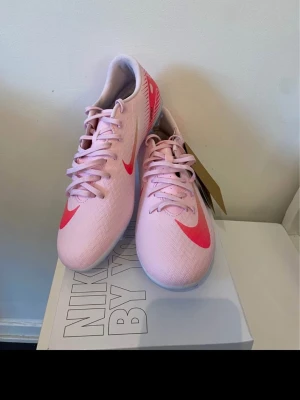 Nike rosa fotbollsskor med röd swoosh - Säljer ett par fotbollsskor från Nike i ljusrosa med röd swoosh på sidan. Skorna har snörning och en platt sula med dobbar för bra grepp på planen. Ovandelen är i syntetmaterial med subtilt mönster och lågt skaft. Perfekt för dig som vill sticka ut på fotbollsplanen.