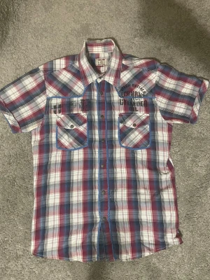 Rutig skjorta från Jack & Jones M - Säljer en rutig skjorta från Jack & Jones i storlek M men kan passa S. Skjortan har korta ärmar, två bröstfickor med knappar och tryckdetaljer fram och bak. Färgerna är röd, blå, vit och svart med blå kant runt fickorna. Materialet är mjuk bomull.