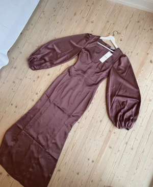 Lång lila satin-klänning från Adoore - Supersnygg långklänning i lila satin från Adoore med v-ringad hals och puffiga långärmar. Klänningen har en figurnära passform och markerad midja som ger en elegant siluett. Perfekt för dig som vill sticka ut med stilrena detaljer och lyxigt material.