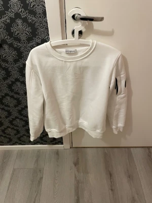 Vit sweatshirt från C.P. Company - Säljer en clean vit sweatshirt från C.P. Company med rund hals och ribbade muddar. Tröjan har en cool svart linsdetalj på vänster ärm som ger en unik touch. Perfekt för dig som gillar stilrena och moderna plagg. Väldigt bra skick! Passar till nästan alla stilar, storlek S