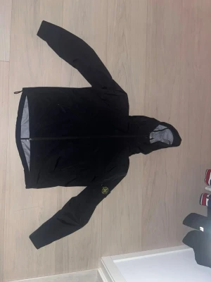 Svart vindjacka från Stone Island - Svart vindjacka från Stone Island med huva och dragkedja framtill. Jackan har ett ikoniskt märke på ärmen och är tillverkad i ett tekniskt, vatten- och vindtätt syntetmaterial med grått foder. Perfekt för dig som gillar streetwear och vill ha en clean look.