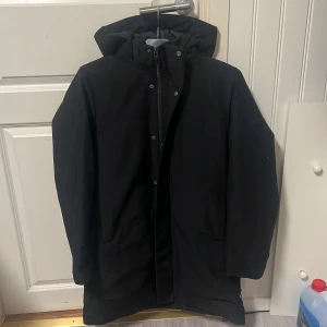 Svart parkajacka från Uniqlo - Säljer en stilren svart parkajacka från Uniqlo. Jackan har en praktisk dragkedja och knappar framtill samt en skyddande huva. Perfekt för kyligare dagar. Nypris 1400kr mitt pris 500. Inte använt den mer än 2 ggr för fick som present men inte min stil