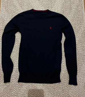 Mörkblå stickad tröja från Polo Ralph Lauren - Snygg mörkblå stickad tröja från Polo Ralph Lauren med klassisk röd logga broderad på bröstet. Tröjan har rund hals, långa ärmar och ribbade muddar vid ärmslut och nederkant. Den har även ett hål vid höger armhåla men går att sy ihop, också två minimala hål i nacken där lappen låg förut men de är mindre än 1 cm ( du kan se de på bilderna) Perfekt för dig som gillar stilrena och tidlösa plagg och har storlek xs-s