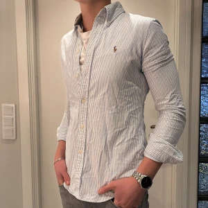 Randig skjorta från Polo Ralph Lauren - Säljer en fräsch vit och blå randig skjorta från Polo Ralph Lauren i custom fit. Skjortan har klassisk krage, knappar framtill och långärmade ärmar. Perfekt för dig som gillar stilrena och tidlösa plagg med snygg passform. Använd 1 gång