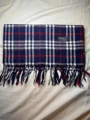 Burberry Halsduk Mörkblå - Säljer en klassisk rutig halsduk från Burberry i mörkblå, vit och röd. Halsduken är tillverkad i 100% lammull och har fransar längs nederkanten. Perfekt för dig som gillar tidlös stil och vill ha något som sticker ut lite extra.