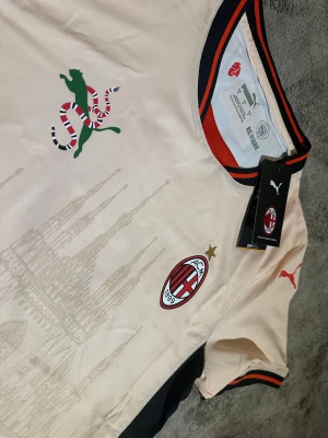 AC Milan Puma matchtröja  - AC Milan matchtröja från Puma i beige med svarta och röda detaljer. Tröjan har klubbmärke, Puma-logga och ett stilrent tryck av Duomo di Milano på framsidan. Rund hals med röd och svart kant. Tillverkad i polyester med Drycell-teknologi.