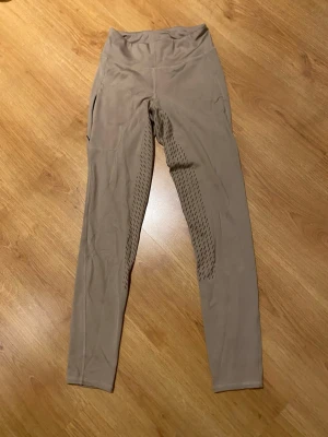 Beige leggings från Domino, strl 36 - Snygga beige leggings från Domino i storlek 36. De har hög midja och är tillverkade av polyamid, polyester och elastan för en stretchig och bekväm passform. Sidorna har diskreta mönsterdetaljer som ger extra stil. Perfekta för träning eller chill.