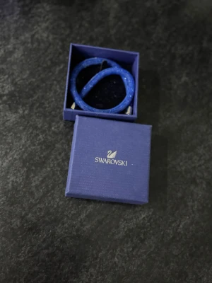 Blå glittrig Swarovski armband - Säljer ett snyggt blått armband från Swarovski med glittrande detaljer och silverfärgat magnetlås. Armbandet har en rund form och är täckt av ett nätliknande material med små paljetter som ger extra bling. Perfekt accessoar för att lyfta din outfit.