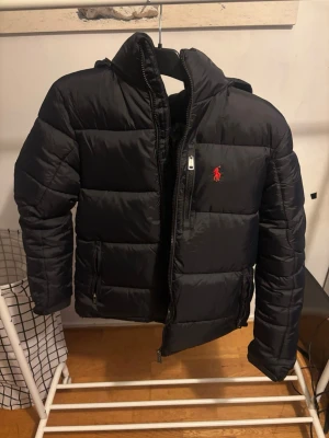 Svart pufferjacka från Polo Ralph Lauren - Svart pufferjacka från Polo Ralph Lauren med röd broderad logga på bröstet. Jackan har huva, två sidofickor med dragkedja och RL-detaljer på zipparna. Perfekt för kalla dagar med sin fluffiga och stilrena look.