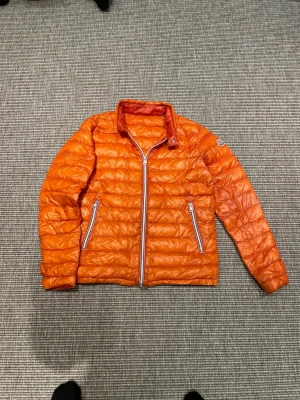 Moncler rigel jacka - Hej, Säljer en orange dunjacka från Moncler med quiltad design och två fickor med dragkedja. Jackan har hög krage med knapp och Moncler-logga på ärmen. Perfekt för dig som vill sticka ut med färg och stil under kyliga dagar. Priset ej hugget i sten!