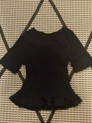 Svart blus med volang från Gina Tricot - Säljer en svart blus från Gina Tricot i storlek 170. Toppen har korta ärmar, smockad midja och en söt volang längst ner. Materialet är mjukt och stretchigt, perfekt för dig som gillar stilrena och feminina detaljer.