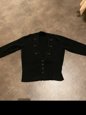 Svart kofta med knappar och v-ringning - Snygg svart kofta med djup v-ringning och dekorativa knappar längs både halsringningen och framtill. Modellen är långärmad och har en ribbad nederkant som ger en schysst passform. Perfekt att slänga över en topp för en chill look.