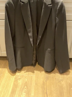 Svart kavaj från C.RO - Stilren svart kavaj från C.RO med klassisk passform och snygga detaljer som knappar vid ärmslut och diskreta fickor fram. Materialet känns mjukt och har en lätt glansig finish. Perfekt för dig som vill ha en clean look med lite edge.