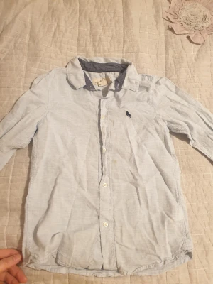 Ljusblå skjorta från L.O.G.G. - Ljusblå skjorta från L.O.G.G. med klassisk krage och kontrastfärg på insidan av kragen. Skjortan har långa ärmar, vita knappar framtill och ett litet broderat motiv på bröstet. Perfekt för dig som gillar enkel och stilren look.