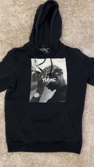 Svart Tupac hoodie med tryck - Svart hoodie med stor Tupac-print på bröstet. Tröjan har huva med snörning och en klassisk känguruficka framtill. Materialet är mjuk bomull och polyesterblandning, perfekt för dig som gillar streetwear och ikoniska motiv.
