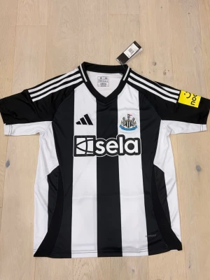 Newcastle adidas fotbollströja - Säljer en officiell Newcastle United fotbollströja från adidas i storlek M. Tröjan har klassiska svarta och vita ränder, klubbmärke på bröstet och sponsortryck. Tillverkad i lätt polyester med AEROREADY-teknologi och slim fit passform. Perfekt för dig som älskar fotboll och snygg design.