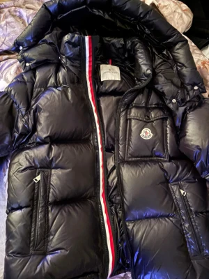 Svart pufferjacka från Moncler - Säljer en svart pufferjacka från Moncler med glansig finish och klassisk logga på bröstet. Jackan har huva, flera fickor och en snygg dragkedja med röd och vit rand längs kanten. Perfekt för kalla dagar och riktigt stilren, storlek 3.