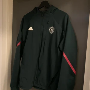 Manchester United hoodie Adidas M - Snygg mörkgrön Manchester United vindjacka från Adidas med huva och dragkedja. Jackan har röda detaljer på ärmarna och klubbens logga på bröstet. Perfekt för dig som vill representera ditt lag och hålla dig bekväm. Passar dig som gillar sportig stil.
