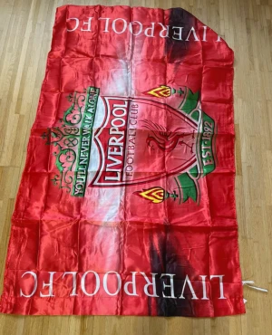 Röd Liverpool FC väst i polyester - Säljer en röd väst med Liverpool FCs ikoniska klubbmärke och texten 'You’ll Never Walk Alone' på framsidan. Västen har en glansig finish och är tillverkad i polyester, med vita och gröna detaljer samt tryckta emblem. Perfekt för dig som älskar fotboll och vill visa ditt stöd.