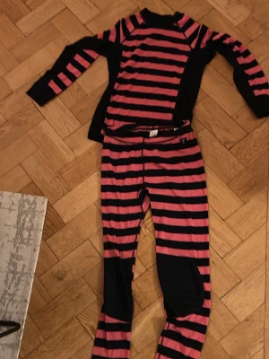 Underställ Polarn O. Pyret 146/156 - Underställ i svart och rosa från Polarn O. Pyret. Setet består av långärmad tröja och byxor med breda ränder och förstärkta knän. Perfekt för kalla dagar och aktiva utomhuslekar. Mjukt material som känns skönt mot huden.