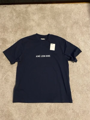 Mörkblå Aimé Leon Dore t-shirt - Snygg mörkblå t-shirt från Aimé Leon Dore med vit logotyp tryckt på bröstet. Klassisk rund hals och korta ärmar. Tillverkad i 100% bomull för skön och mjuk känsla. Perfekt för dig som gillar stilrena och cleana plagg med street-vibe.