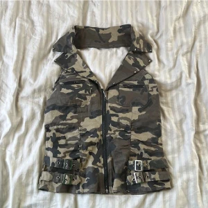 Y2k Camo väst/topp med dragkedja från Lydia - Snygg camouflageväst från Lydia med coola detaljer som stora kragar, dragkedja framtill, två bröstfickor med zip och dubbla bältesremmar i midjan. Västen är i bomull med lite stretch för skön passform. Perfekt för dig som vill sticka ut.  Perfekt till sommaren.  Den är stretchig.  Och den har padding vid brösten (se sista bilden) passar bäst om du har stolek L eller M (exempel 90E är för stort). Bild 2 och 3 är en annan tröja, är för att vissa hur toppen sitter på