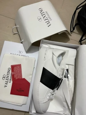 Valentino Garavani Open sneakers - Säljer ett par snygga Valentino Garavani Open sneakers i vitt skinn med en bred svart läderdetalj över mitten. Klassisk rund tå, platt sula och vita snören. Kommer med originalkartong och dustbag. Perfekt för dig som gillar lyxiga och stilrena sneakers.