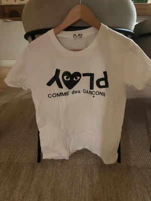 Vit Comme des Garçons Play t-shirt - Vit t-shirt från Comme des Garçons Play med svart tryck och det ikoniska hjärtat med ögon på bröstet. Klassisk passform med rund hals och korta ärmar. Perfekt för dig som gillar streetwear och stilrena plagg.