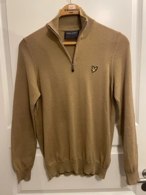 Beige stickad half zip från Lyle & Scott - Snygg beige stickad tröja från Lyle & Scott med half zip och lång ärm. Tröjan har en liten broderad gul fågel på bröstet och ribbade muddar vid ärmslut och nederkant. Perfekt för dig som gillar klassisk stil med en modern touch.