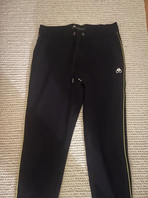 Svarta mjukisbyxor från Moose Knuckles - Svarta mjukisbyxor från Moose Knuckles med gul piping längs sidorna och logga på benet. Byxorna har snörning i midjan och är gjorda i mjukt bomullsmaterial. Perfekta för chill eller träning, med en clean och sportig vibe.