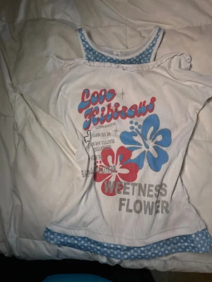 Cute core topp - Säljer en vit topp med offshoulder och blått prickigt linne under. Framsidan har tryck med stora hibiskusblommor i blått och rosa samt text.  