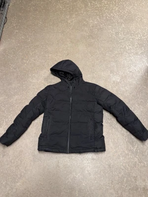 Svart pufferjacka från H&M Sport - Svart pufferjacka från H&M Sport med huva och dragkedja framtill. Jackan har två sidofickor med dragkedja och är tillverkad i polyester. Perfekt för kalla dagar och har en stilren look som funkar till många outfits.