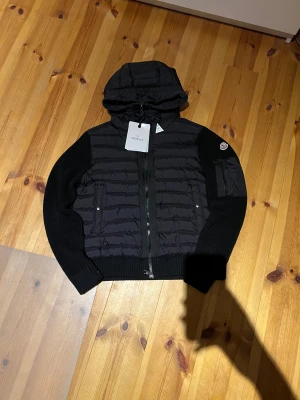 Moncler cardigan - BILLIGARE VID SMIDIG AFFÄR. Säljer då jag är sugen på nytt. Svart cardigan från Moncler med huva och stickade ärmar samt baksida. Dragkedja framtill, ribbade muddar och en ficka med dragkedja på ärmen. Klassisk Moncler-logga på vänster ärm och quiltad kropp. Perfekt för dig som vill ha en snygg och varm cardigan. Den är även väldigt varm, har använt den med T-shirt under i vinter och har inte frusit alls.