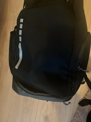 Svart Nike ryggsäck Elite - Säljer en svart Nike ryggsäck med Elite-logga och silvriga detaljer. Väskan har flera fack med dragkedja, meshficka på insidan och justerbara axelremmar. Perfekt för dig som vill ha en sportig och funktionell väska med stilren design.