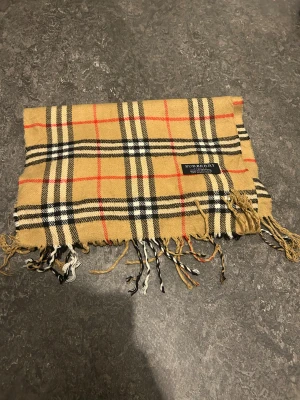 Burberry halsduk  - Säljer en klassisk rutig halsduk från Burberry i beige, svart, vit och röd. Halsduken har fransar i kanterna och den ikoniska Burberry-taggen. Perfekt accessoar för att lyfta din outfit har ingen användning 