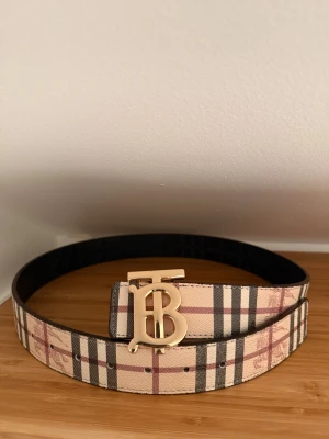  Burberry bälte - Säljer ett snyggt Burberry bälte med klassiskt rutigt mönster i beige, svart och röd. Bältet har ett stort guldfärgat spänne med Burberrys ikoniska TB-logga. 