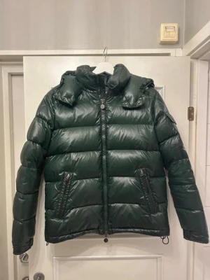 Mörkgrön Moncler pufferjacka - Säljer en mörkgrön pufferjacka från Moncler med hög krage och avtagbar huva. Jackan har två snedställda fickor med dragkedja, Moncler-logga på ärmen och quiltad design. Materialet är glansig polyester och dunfyllning, perfekt för kalla dagar. MÖTS BARA ‼️
