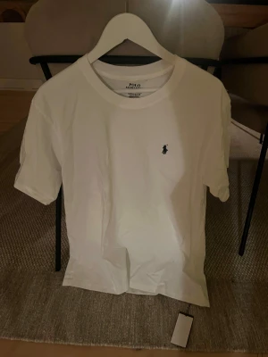 Vit t-shirt från Polo Ralph Lauren - Vit t-shirt från Polo Ralph Lauren med klassisk rund hals och korta ärmar. T-shirten har en liten broderad logga på bröstet och är gjord i mjuk bomull. Enkel och stilren design som funkar till många olika looks.