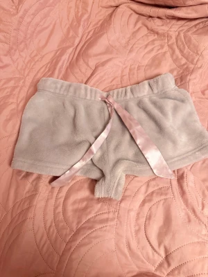 Ljusrosa shorts  - Supermjuka grå shorts i plysch med söt kattansikte och öron på baksidan samt texten 'MEOOOW'. Framtill finns en ljusrosa satinrosett i midjan. 